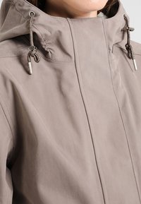 Giacca con cappuccio grigio chiaro caratterizzata da una texture liscia, coulisse regolabili e design minimalista con sottili dettagli di cucitura.