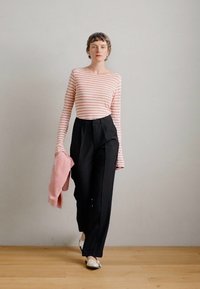 Blusa de mangas longas às riscas em rosa claro e branco, calças pretas de cintura alta, segurando um suéter texturizado rosa e usando sapatos com detalhes brancos.