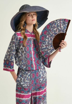 Donna che indossa un abito a fantasia rosa e blu, con un grande cappello da sole blu, che tiene un ventaglio decorativo abbinato, su uno sfondo uniforme.