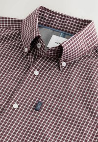 Chemise à carreaux bordeaux et blanche avec boutons blancs et un petit logo brodé sur la poitrine.