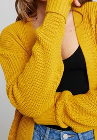 Cárdigan amarillo de punto con un patrón texturizado y puños de canalé, usado sobre una blusa negra, parcialmente visible. Estilo casual, corte relajado.