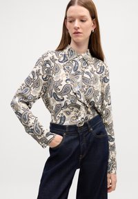 Long-sleeve blouse in cream with blue paisley pattern, button-down collar, and a tied waist detail, paired with dark denim high-waisted trousers.