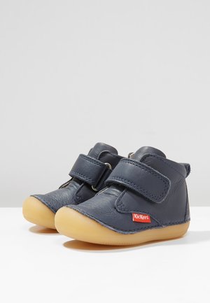 Par navyblå lædersko til småbørn med beige gummisåler og velcroremme, med et rødt Kickers-mærke på siden.