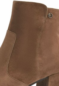 Bottine en daim marron avec un design épuré, un panneau latéral incurvé et des accents en métal argenté. Surface texturée et talon robuste.