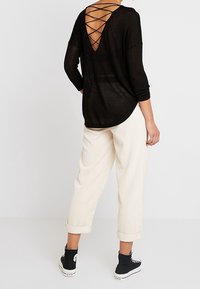 Schwarzes Strickoberteil mit einem tiefen V-Ausschnitt und gekreuzten Details, kombiniert mit cremefarbenen Cordhosen und schwarzen High-Top-Sneakern.