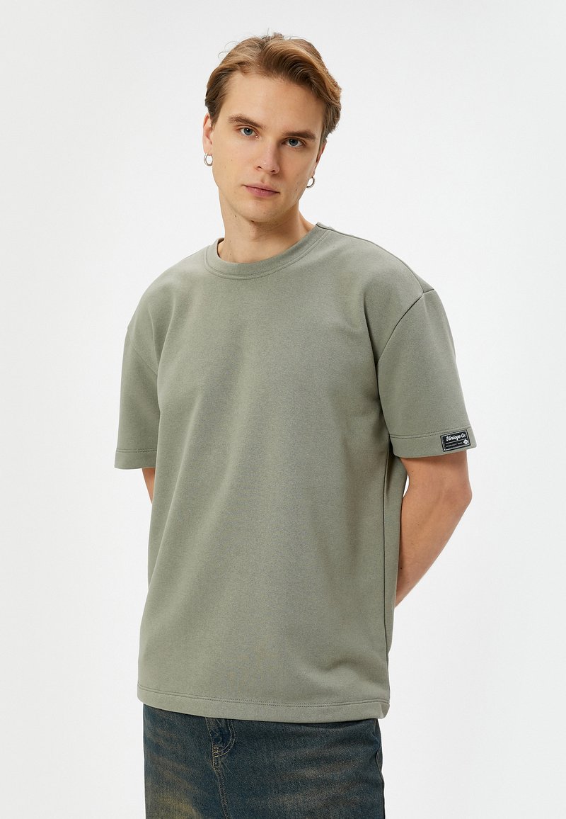 Camiseta de algodón verde claro con mangas cortas, un cuello redondo y una pequeña etiqueta de marca en la manga izquierda. Textura suave, corte casual.