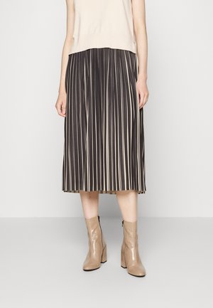 Frau trägt ein beiges ärmelloses Oberteil, einen schwarz-beigen Midi-Rock mit vertikalen Streifen und beige Lackleder-Stiefeletten mit Blockabsätzen.