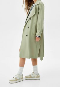 Jasnozielony trench z klapami, podwójnym zapięciem z przodu, bocznymi kieszeniami i paskiem do przewiązywania, w połączeniu z białymi sneakersami i teksturowanymi skarpetami.