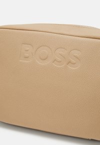 Beige faux läderväska med ett präglat "BOSS"-logotyp. Har en slät yta och rundad form med dragkedjeöppning.