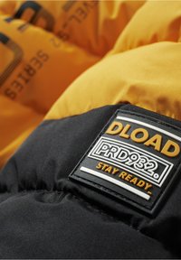 Doudoune avec une partie inférieure noire et une partie supérieure jaune, arborant une patch noir texturé avec l'inscription "DLOAD PRD932 STAY READY."