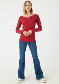 Suéter rojo de punto con un amplio escote en V, puños y dobladillo acanalados. Combinado con jeans acampanados azules y zapatillas beige. El modelo está de pie con las manos entrelazadas.