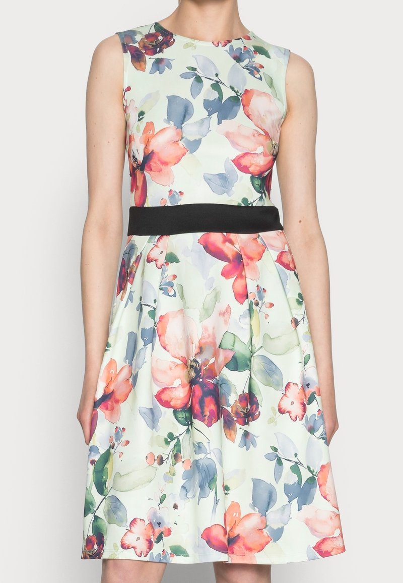 Robe blanche sans manches avec un grand imprimé floral rose, rouge et bleu et une ceinture noire, portée par une personne debout.