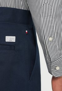 Granatowe spodnie z boczną kieszenią, na której znajduje się biały metka z napisem "HILFIGER". Częściowo widoczny jest rękaw czarno-białej w paski koszuli.