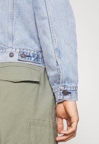 Ljusblå jeansjacka med knäppt manschett; tillsammans med olivgröna, strukturerade cargo-byxor med en ficka med lock.