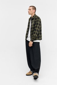 Camisa de flanela xadrez verde e preta, com uma camisa branca por baixo, combinada com calças largas pretas e sapatos bege. Design simples e ajuste casual.