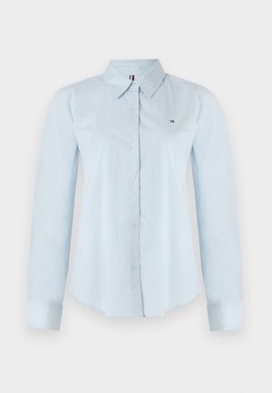 Chemise à manches longues bleu clair avec un bouton sur le devant et un col. Présente une broderie de logo discrète et une texture de tissu lisse.