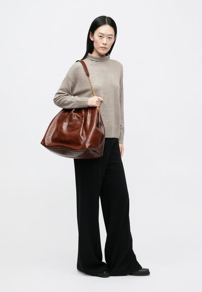 Καφέ δερμάτινη τσάντα ώμου με χαλαρή φόρμα, χρυσή αλυσίδα για λουρί και λείο υλικό, κρατιέται από μοντέλο που φοράει μπεζ ζιβάγκο και μαύρο παντελόνι.