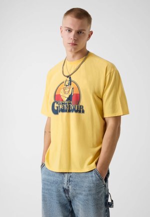 Junger Mann mit kurzen Haaren, der ein gelbes T-Shirt mit Grafik und hellblaue Jeans trägt, steht mit den Händen in den Taschen vor weißem Hintergrund.