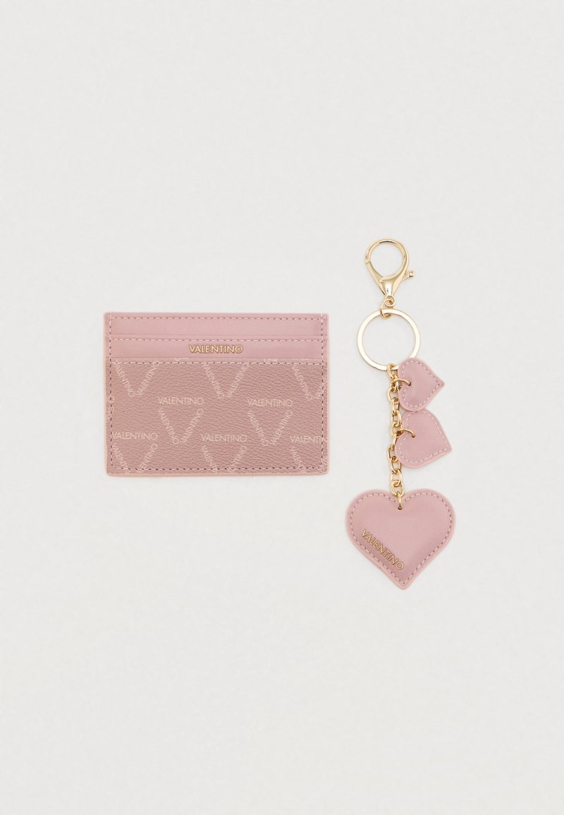 Ροζ δερμάτινη θήκη καρτών με ανάγλυφο μοτίβο λογότυπου, εφοδιασμένη με χρυσό κλειδοκράτη με δύο ροζ καρδιές. Λείο υλικό, συμπαγής σχεδίαση.