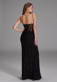 Femme portant une robe longue noire à sequins avec un dos transparent à fines bretelles, debout avec une main sur la hanche devant un fond gris.
