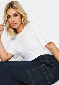 Biała bawełniana koszulka z krótkim rękawem, w połączeniu z ciemnoniebieskimi spodniami cargo z kieszeniami naszytymi i kontrastowym szyciem. Model siedzi w swobodnej pozycji.