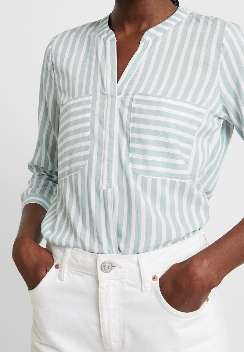 Blouse à rayures bleu clair en tissu doux, avec un col mao, deux poches poitrine, coupe décontractée, portée avec un pantalon blanc.