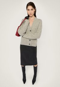 Szary kardigan w warkocz o splocie styrdowym z trzema guzikami, zestawiony z czarną spódnicą ołówkową i czarnymi botkami za kolano, trzymający małą czerwoną torebkę.