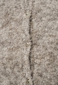 Καρδιάκα σε καφέ και μπεζ υφή με χρυσά κουμπιά κατά μήκος του μπροστινού μέρους, κατασκευασμένη από μαλακό, αφράτο πλεκτό υλικό.