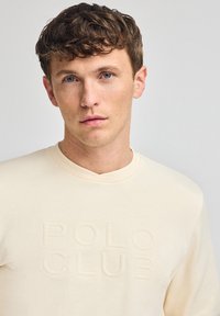 Felpa beige con collo a giro, caratterizzata da un logo in rilievo "POLO CLUB". Realizzata in tessuto morbido, con una texture liscia e un design casual.