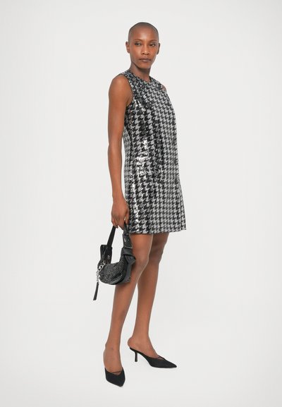 Φόρεμα με παγιέτες σε καρό σχέδιο σε μαύρο και ασημί, χωρίς μανίκια, σε σχήμα Α, συνδυασμένο με μια μικρή μαύρη τσάντα και ψηλοτάκουνα παπούτσια.