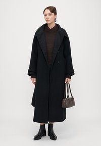 Melna vilna mēteli ar lielām apmalēm un divrindu pogām, uzvilkts virs brūnas adītas džempera. Ietver mazu pelēku rokassomi un potīšu zābakus.