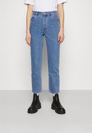 Persoon draagt blauwe, rechte jeans met hoge taille, wit shirt, witte sokken en zwarte, stevige enkellaarsjes tegen een effen achtergrond.