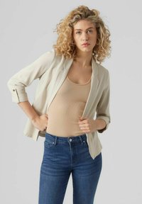 Beige sako s měkkou texturou, přizpůsobený střih a ohrnuté rukávy, spárované s béžovým vrškem s žebrovaným vzorem a tmavě modrými džíny.
