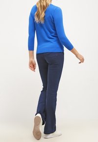 Blå stickad tröja med tre fjärdedels ärmar, kombinerad med mörka flarejeans och vita sneakers, sedd bakifrån.