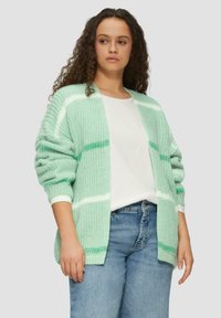 Hellgrüner Cardigan mit weißen und dunkleren grünen Streifen. Stricktextur, offen vorne, herunterhängende Schultern, getragen über einem weißen Hemd und Jeans.