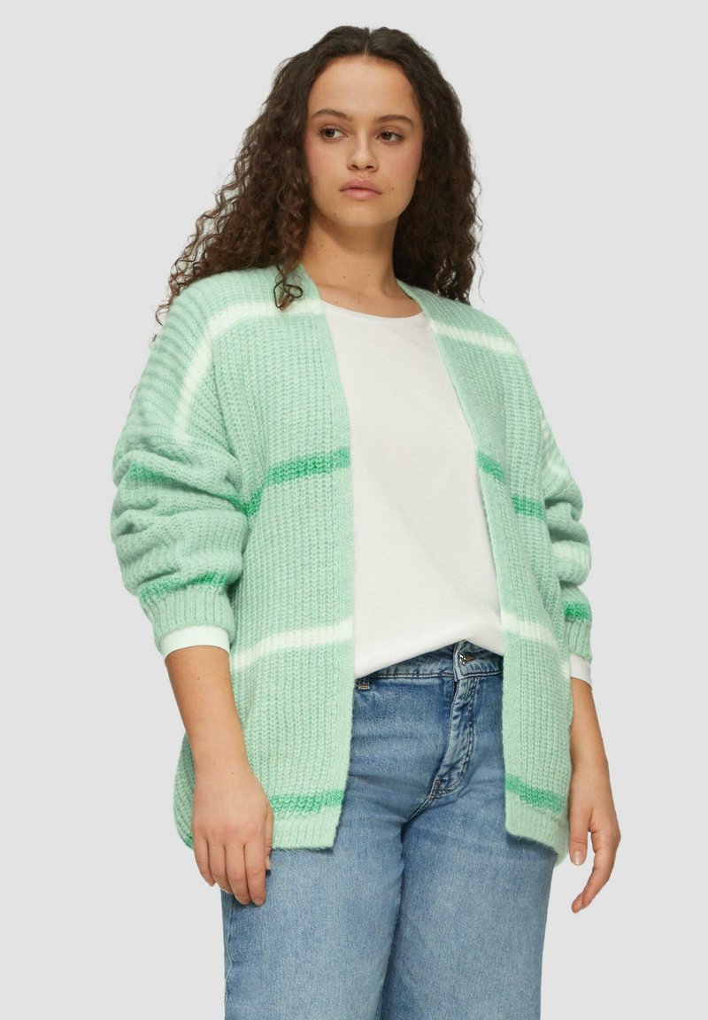 Hellgrüner Cardigan mit weißen und dunkleren grünen Streifen. Stricktextur, offen vorne, herunterhängende Schultern, getragen über einem weißen Hemd und Jeans.
