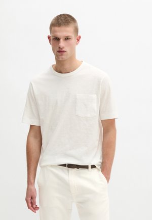 Junger Mann trägt ein weißes Kurzarmhemd mit Brusttasche und weiße Hose, steht mit einer Hand in der Tasche.