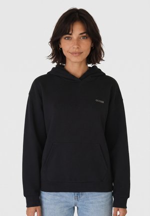 Jeune femme aux cheveux courts et foncés portant un sweat à capuche noir Guess et un jean bleu clair sur un fond clair uni.