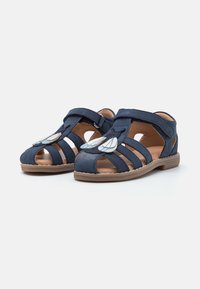 Sandalias azul marino con diseño recortado, que cuentan con una correa de velcro y detalles decorativos en azul claro. Hechas de ante con una suela marrón.