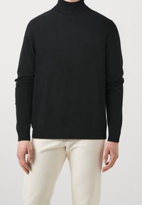 Mann in einem schwarzen langärmeligen Rollkragenpullover und cremefarbenen Hosen, der vor einem schlichten hellen Hintergrund steht.