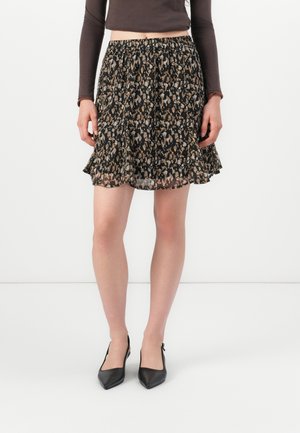 Nainen, jolla on lyhyt musta ja ruskea leopardikuosinen hame, pitkähihaiset ruskeat yläosa ja mustat teräväkärkiset slingback-korkokengät, seisoo valkoisella lattialla.