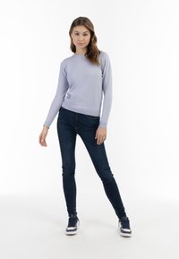 Pulover lung cu mânecă lungă de culoare lavandă deschisă, cu manșete cu nervuri, asortat cu blugi skinny de culoare albastru închis și adidași bleumarin, pe un fundal simplu.