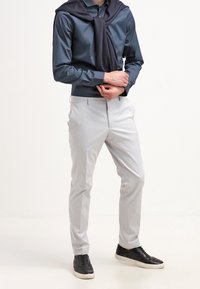 Námořnická košile na knoflíky, světle šedé chinos kalhoty a černé tenisky. Tmavý svetr přehozený přes ramena. Hladká látka a slim fit.