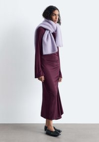 Langes kastanienbraunes Kleid mit gerafftem Seitendesign, einem weichen, helllila Schal und schwarzen Slippern. Glatte Textur und figurbetonte Silhouette.