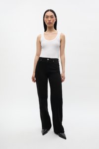 Jeans neri a vita alta con design a gamba dritta, abbinati a un top bianco aderente. La modella indossa scarpe nere a punta. Sfondo minimalista.
