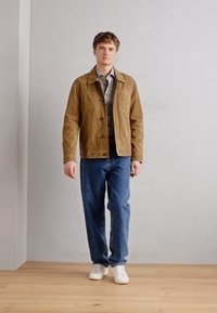 Brown suede jacket over a patterned shirt, paired with blue denim jeans and white sneakers. Neutral backdrop and wooden flooring.