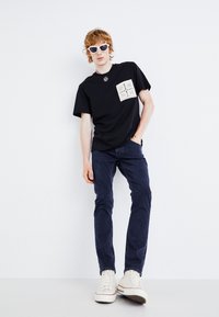 T-shirt noire avec une poche à motif blanc, associée à un jean bleu foncé et des baskets blanches. Des lunettes de soleil rondes complètent le look.