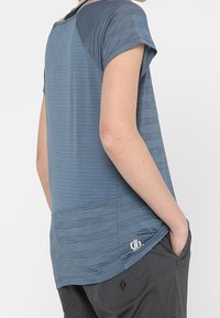 Sininen urheilupaita, jossa on lyhyet hihat, teksturoitu kuvio ja pidempi selkähelmassa. Pieni logo alaosassa sivulla.