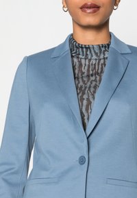 Světle modrý blazer s jedním zapínáním na knoflík, klopovými límci a předními kapsami, přes vzorovaný černý a modrý top.
