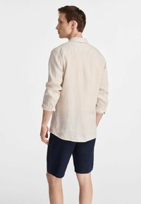 Męska jasnobeżowa koszula lniana z podwiniętymi rękawami, karczkiem z tyłu i luźnym krojem, w zestawieniu z granatowymi szortami. Minimalistyczny design, styl casual.